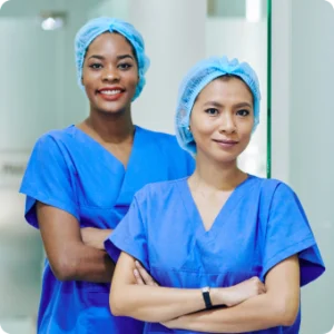 Two women wearing scrub suits stand together, smiling in a healthcare setting.