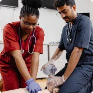 Two nurses in scrubs practicing medical procedures on a training dummy in a clinical setting.