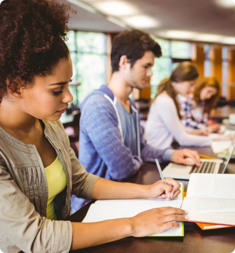 A group of diverse students studying together at tables in a library, surrounded by books and study materials.