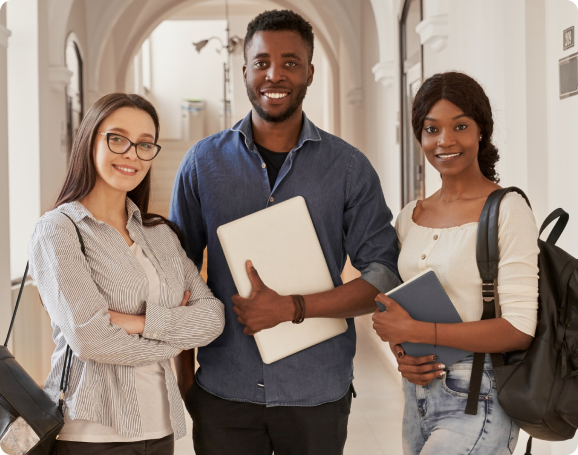 Three students stand in a hallway, one holding a laptop, engaged in conversation and smiling at each other.