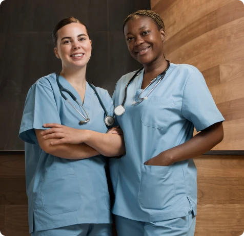 Two female nurses stand together in front of a wall, wearing scrubs and smiling at the camera.